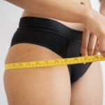 A woman in black panties measuring her hips with yellow measuring tape.